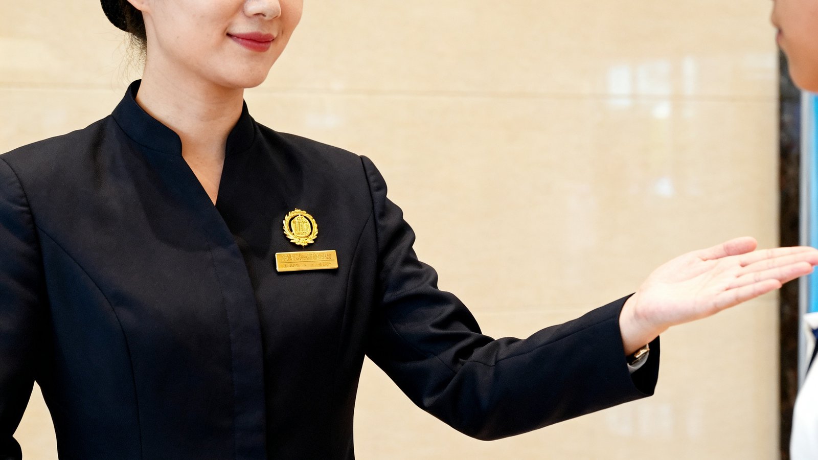 一位仪态端庄的女性礼宾经理正在模拟接待大厅内指导年轻礼宾人员练习引导手势，她身着深色制服，胸前佩戴金色公司徽章，表情专注而温和
